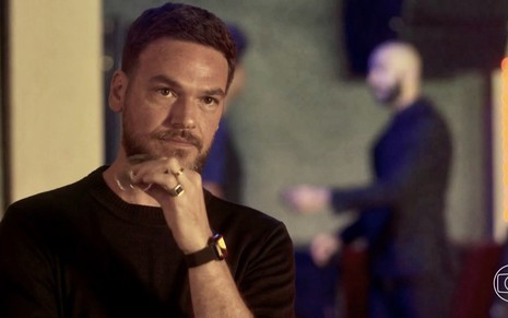 O ator Emilio Dantas usa camiseta preta e está com as mão no queixo em cena da novela Vai na Fé como Theo