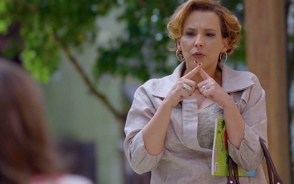 A atriz Ana Beatriz Nogueira como Eva está em uma praça, ela cruza os dedos em um xis na frente da boca, como se jurasse segredo, em cena de A Vida da Gente