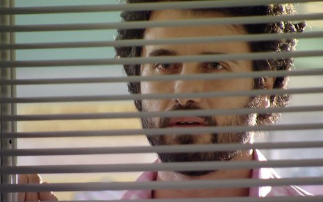 O ator Eriberto Leão como Gabriel está do lado de fora de uma janela com uma persiana entre-aberta em cena de A Vida da Gente
