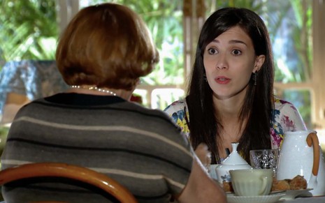 A atriz Nicette Bruno como Iná, de costas, está sentada a uma mesa de café da tarde e olha para Marjorie Estiano, a Manuela, que está em sua frente à direita em cena de A Vida da Gente
