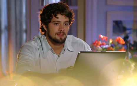 O ator Rafael Cardoso caracterizado como Rodrigo está sentado à mesa em frente a um laptop prateado em cena de A Vida da Gente