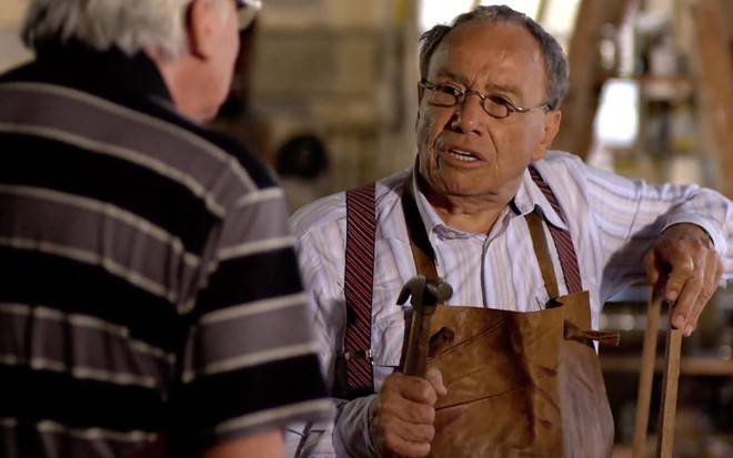 O ato Stênio Garcia como Laudelino está com o braço direito apoiado em uma estrutura de madeira e segura um martelo com a mão direita em cena de A Vida da Gente