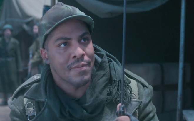 Sentado em acampamento, Bento (Matheus Dias) está com uniforme de soldado em cena de Além da Ilusão