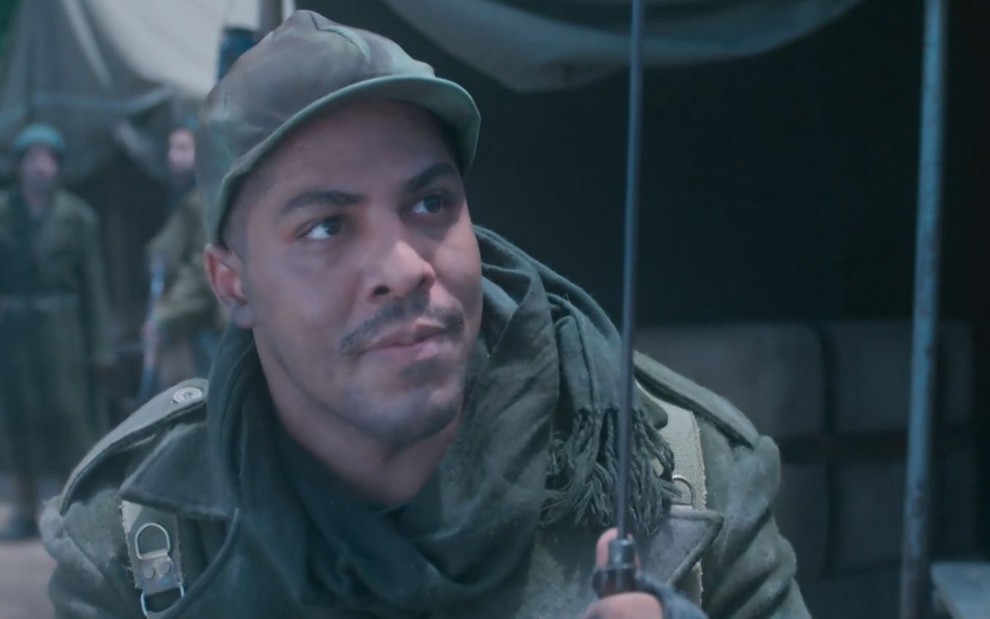 Sentado em acampamento, Bento (Matheus Dias) está com uniforme de soldado em cena de Além da Ilusão
