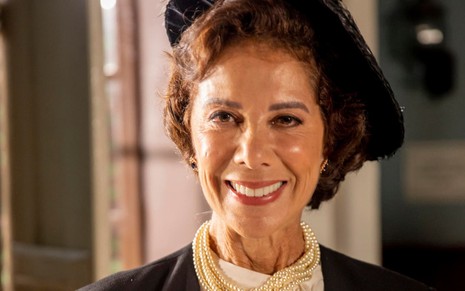 Angela Vieira caracterizada como Lisiê: atriz tem os cabelos presos em um coque e usa um chapéu delicado. Ela também veste um lenço branco, amarrado no pescoço, e um terninho preto enquanto sorri para a câmera