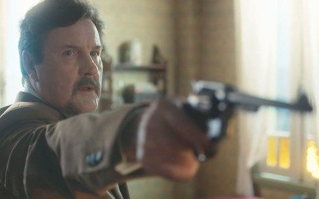 O ator Antonio Calloni como Matias aponta uma arma para a direita dentro do quarto de uma pensão em cena de Além da Ilusão