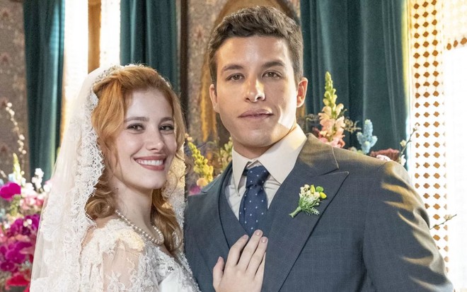 Caroline Dallarosa e Ricky Tavares estão sorridentes para a câmera, com os trajes de noivos