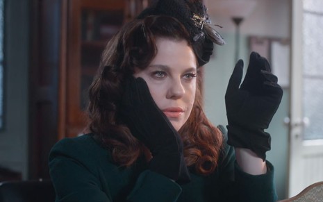 A atriz Bárbara Paz com o braço apoiado em uma mesa e a mão esquerda no rosto como Úrsula, toda de preto, em cena de Além da Ilusão