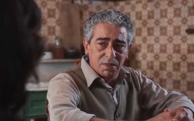 Benê (Claudio Jaborandy) está sentado em mesa e tem expressão de tristeza em cena de Além da Ilusão, novela das seis da Globo
