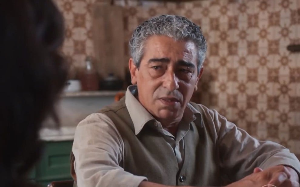 Benê (Claudio Jaborandy) está sentado em mesa e tem expressão de tristeza em cena de Além da Ilusão, novela das seis da Globo