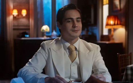 Joaquim (Danilo Mesquita) está sentado em mesa de restaurante e sorri em cena de Além da Ilusão