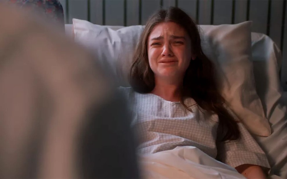 Debora Ozório tem os olhos cheios de lágrimas em cena de Além da Ilusão; a atriz está no leito de um hospital