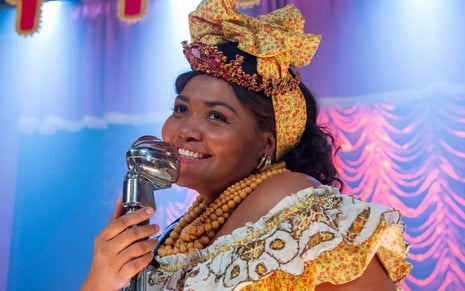 Gaby Amarantos usa um vestido em estilo ciganinha, colares de contas e um laço grande na cabeça; ela segura o microfone enquanto sorri em cena de Além da Ilusão
