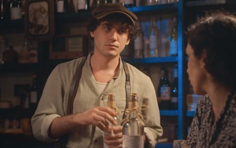 O ator Guilherme Prates como Lorenzo segura uma garrafa de cachaça atrás do balcão de um bar em Além da Ilusão