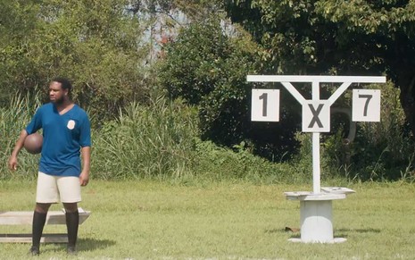 Guilherme Silva, o Onofre, com um uniforme de futebol azul em frente a um placar em que se vê 1 x 7