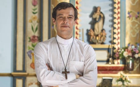 Jayme Matarazzo caracterizado como padre Tenório: ele veste uma bata branca e um crucifixo, e dá um sorriso para a câmera; ao fundo, é possível ver a imagem de uma santa.