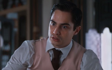 Danilo Mesquita, caracterizado como Joaquim, tem a expressão indignada em cena de Além da Ilusão; ator usa uma camisa branca, uma gravata vermelha e um colete rosa