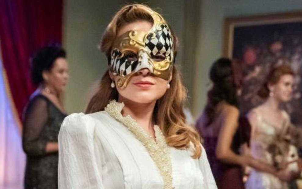 Larissa Manoela, caracterizada como Isadora; ela usa um terninho branco, com detalhes dourados, e uma máscara branca com estampa de losangos pretos e detalhes também dourados em cena de Além da Ilusão