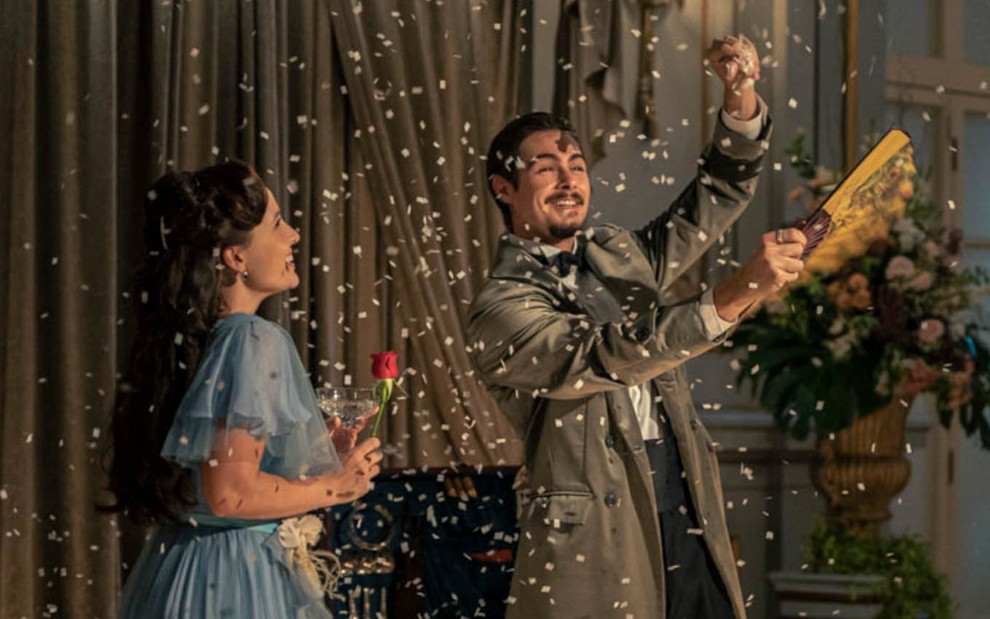 A atriz Larissa Manoela, como Elisa, segura uma rosa com a mão direita enquanto vê Rafael Vitti, o Davi, fazer uma mágica com um leque e confetes em cena de Além da Ilusão