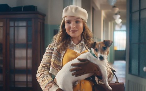 A atriz Larissa Manoela com roupas da década de 1940 segura um cachorro da raça fox paulistina em cena de Além da Ilusão