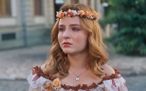 Isadora (Larissa Manoela) chora em cena de Além da Ilusão, novela das seis da Globo, na qual usa adorno de flores e vestido colorido