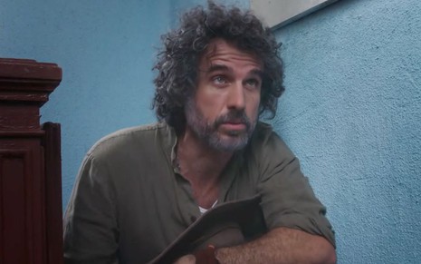 Eriberto Leão em cena de Além da Ilusão: ele está com o cabelo na nuca, camisa verde puída e olha para alguém fora do quadro