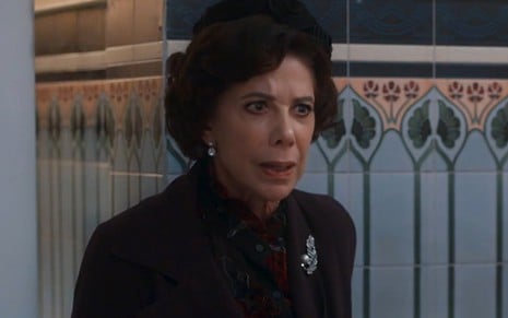 Angela Vieira, caracterizada como Lisiê, tem a expressão chocada em cena de Além da Ilusão; ela veste casaco e chapéu pretos e tem os cabelos presos