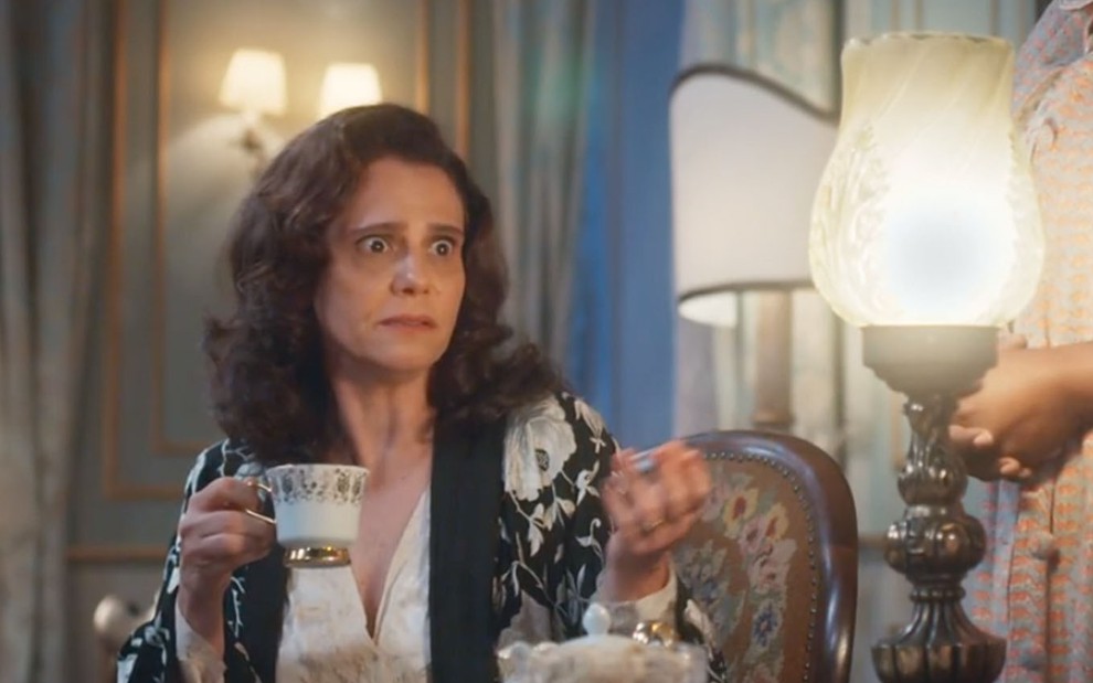 Com expressão de susto, a atriz Malu Galli como Violeta segura uma xícara de chá em cena de Além da Ilusão