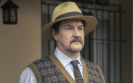 Antonio Calloni, caracterizado como Matias, encara ponto fixo; ele veste chapéu de palha, camisa, gravata e colete de lã em ensaio fotográfico de Além da Ilusão