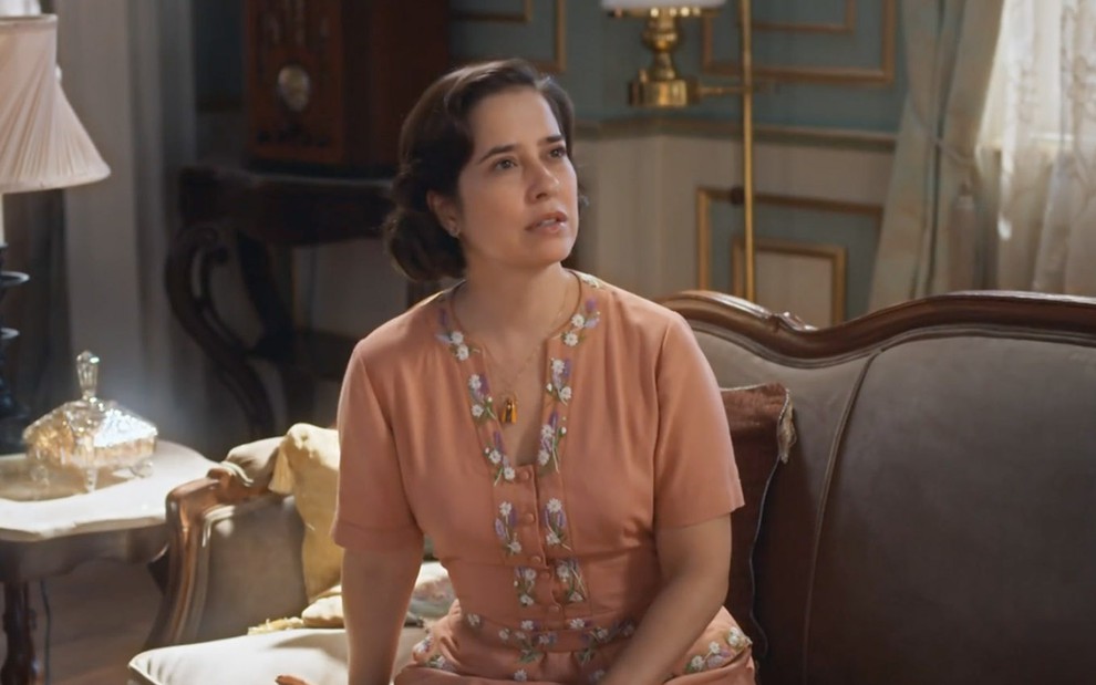 A atriz Paloma Duarte está caracterizada como sua personagem de Além da Ilusão sentada no sofá com uma expressão de susto e surpresa