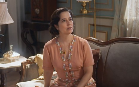 A atriz Paloma Duarte como a Heloísa sentada em um sofá com cara de poucos amigos em cena de Além da Ilusão