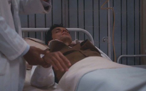 Fabrício Belsoff está deitado em uma cama hospitalar, com os olhos fechados; um médico não identificado segura sua mão inerte em cena de Além da Ilusão