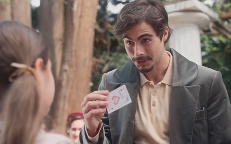 O ator Rafael Vitti como Davi segura um ás de copas em que se lê escrito Isadora em uma praça de Poços de Caldas em cena diurna de Além da Ilusão