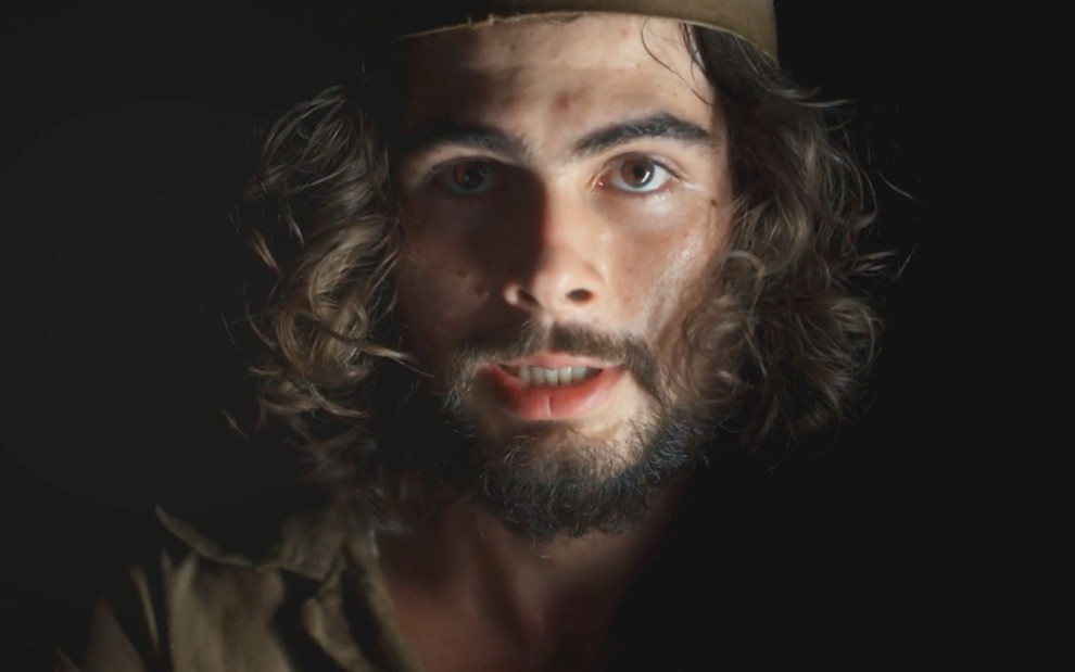 O ator Rafael Vitti com cabelos e barba grandes, com roupas de prisioneiro, em cena de Além da Ilusão