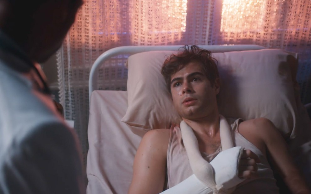 O ator Rafael Vitti como Davi está deitado em um leito de hospital com o braço direito engessado em cena de Além da Ilusão