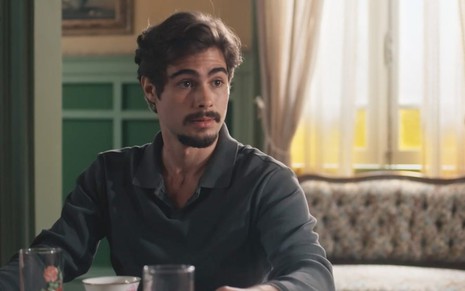 O ator Rafael Vitti, com expressão de surpresa, sentado à mesa como o Davi em cena de Além da Ilusão