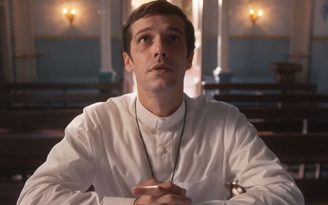 Jayme Matarazzo caracterizado como padre Tenório: ele veste uma camisa branca e um crucifixo. O ator olha para o alto, no meio de uma reza, em cena de Além da Ilusão