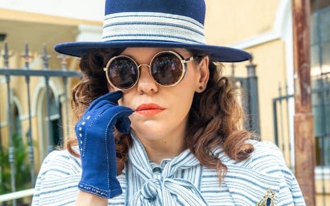 Bárbara Paz caracterizada como Úrsula em Além da Ilusão; atriz usa chapéu e luvas azul royal. Ela tem a expressão séria enquanto encara a câmera em ensaio de divulgação.