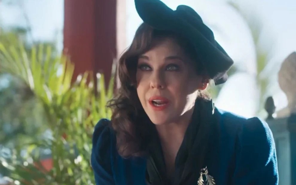 Bárbara Paz caracterizada como Úrsula em Além da Ilusão; atriz usa chapéu e luvas azul royal. Ela tem a expressão séria em cena de Além da Ilusão