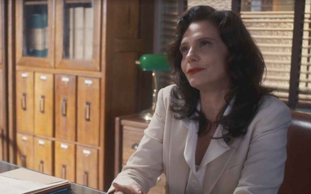 Malu Galli, caracterizada como Violeta, está apoiada sobre a mesa do escritório; ela encara Marcello Novaes, o Eugênio, com deboche em cena de Além da Ilusão