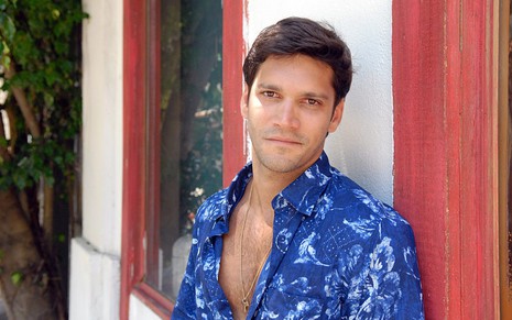 Armando Babaioff posa com camisa azul florida e sorriso tímido, caracterizado como Thales de Ti Ti Ti