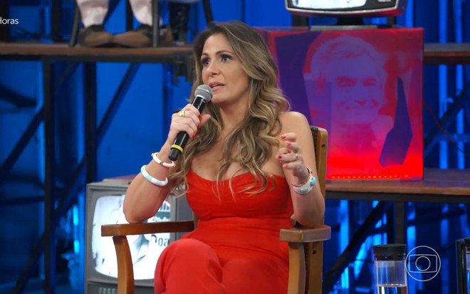 Lega Begliomini, do BBB1, com conjunto vermelho e sentada em cadeira do Altas Horas, com microfone na mão e expressão de pavor