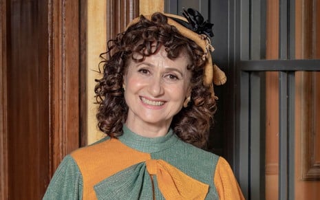 Zezé Polessa caracterizada como Cândida; ela usa um vestido verde a amarelo e sorri para a câmera em ensaio de divulgação de Amor Perfeito