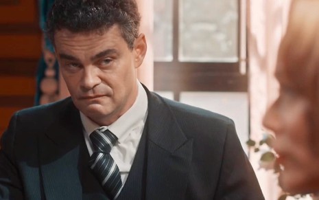 O ator Carmo Dalla Vecchia usa terno e gravata e está com expressão séria em cena de Amor Perfeito como Érico