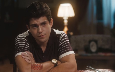 O ator Daniel Rangel está com camisa polo e braços cruzados em cima de uma mesa em cena da novela Amor Perfeito
