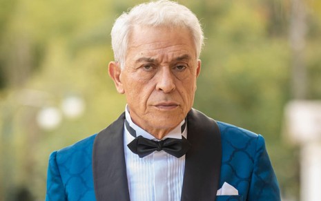 Paulo Gorgulho caracterizado como Leonel em Amor Perfeito, de terno azul, camisa branca e gravata preta, olhar sério