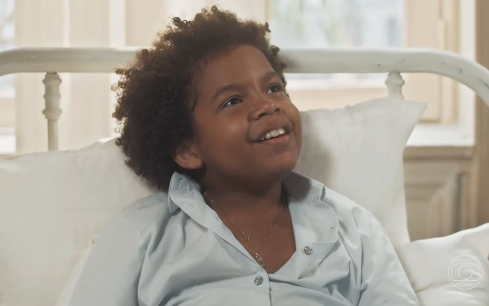 Levi Asaf caracterizado como Marcelino; ele usa um pijama e está deitado numa cama de hospital em cena de Amor Perfeito. O ator dá um leve sorriso.