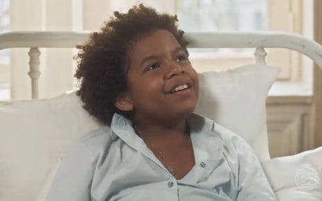 Levi Asaf caracterizado como Marcelino; ele usa um pijama e está deitado numa cama de hospital em cena de Amor Perfeito. O ator dá um leve sorriso.
