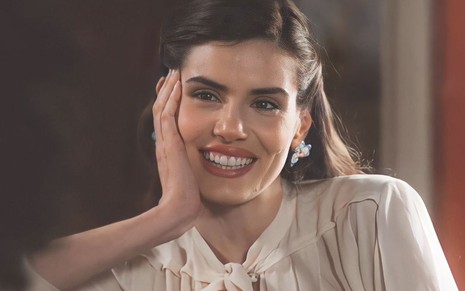 A atriz Camila Queiroz sorri e está com o rosto apoiado em uma de suas mãos em cena de Amor Perfeito como Marê