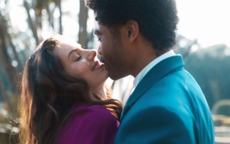 Camila Queiroz beija Diogo Almeida em cena de Amor Perfeito; ela usa um vestido roxo, e ele, um terno azul